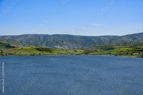Wide angle view of a pristine freshwater reservoir surrounded by lush green hills and craggy peaks. The calm water reflects the blue sky in a tranquil and  remote wilderness. Vast serene lake.
