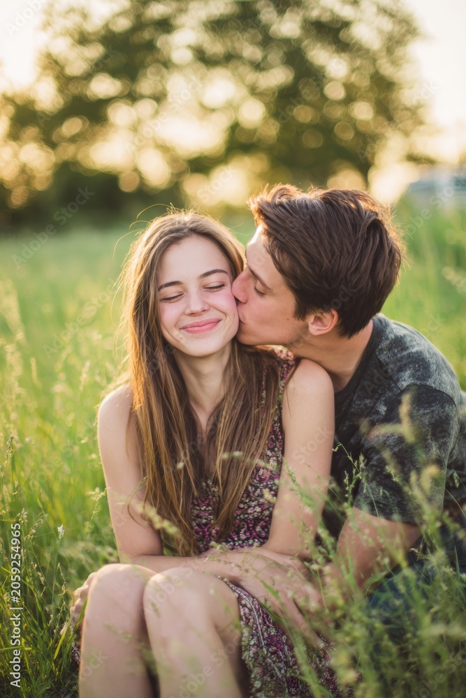 Fototapeta premium Lovely couple romantic moment. A man and woman embracing and kissing in a field of tall grass at sunset. The woman, wearing a dress, has a smile on her face. The background is slightly blurred.