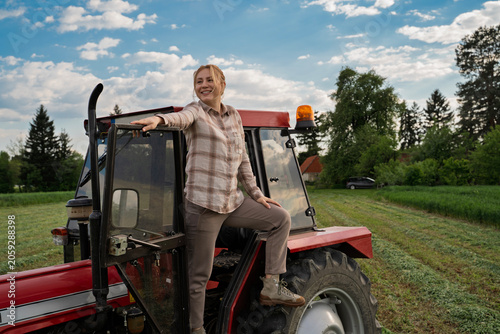 Happy satisfied woman on her land standing on the steps of the tractor and smiling looking ahead 