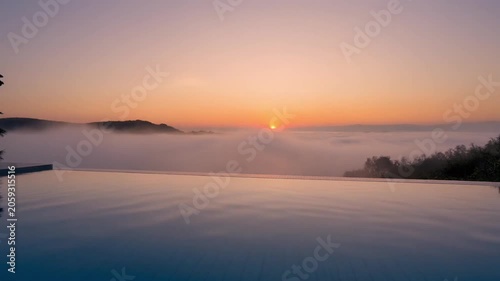 Sunrise over misty valley from infinity pool edge at dawn. Serene dawn view of a golden sunrise over low clouds and hills, seen from a calm infinity pool.