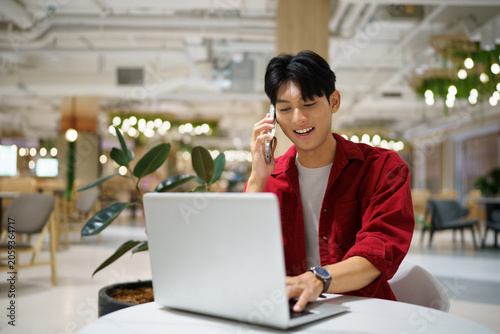 Young Asian Man Talking on Phone While Working on Laptop in Modern Co working Space