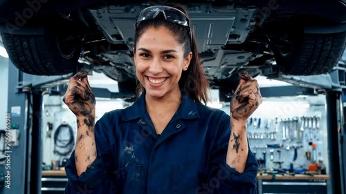 Woman in blue coveralls stands under a lifted car. She holds greasy hands near the vehicle. She smiles confidently. Tools fill the garage background. This shows automotive work
