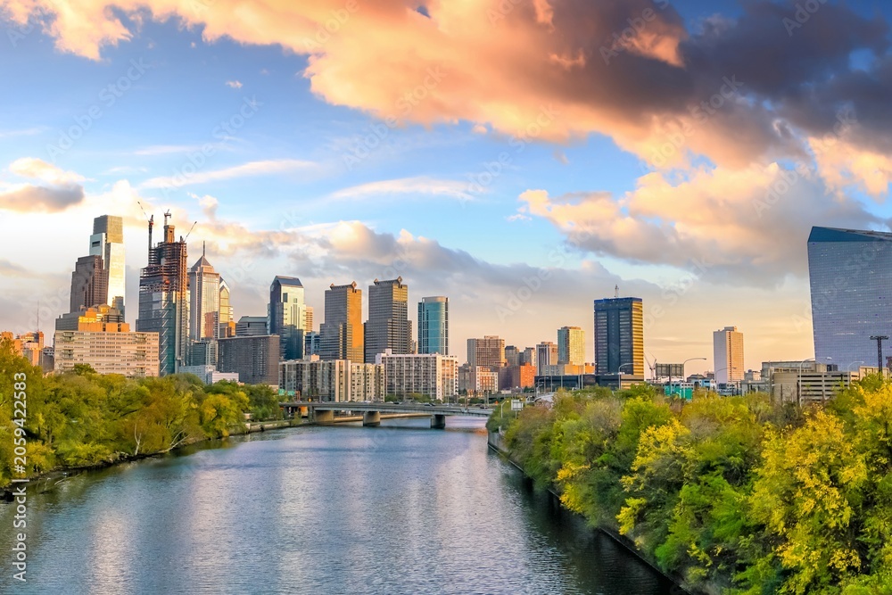 Fototapeta premium Scenic view of the Philadelphia skyline along the Schuylkill River at sunset, featuring modern skyscrapers, vibrant clouds, lush riverside foliage, and reflections in calm water.