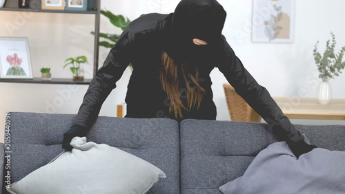Fotografie Masked Burglar Searching Behind Sofa Cushions in Bright Living Room