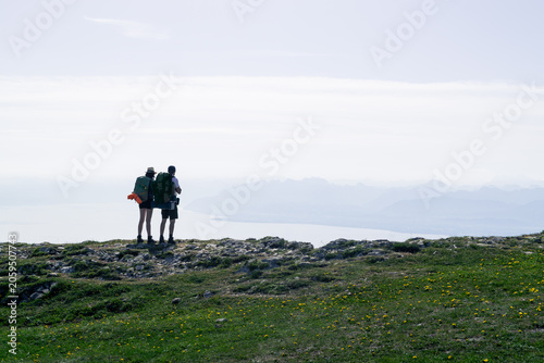 Silhouettes of two hikers with backpacks standing on a rocky alpine meadow overlooking a bright lake and distant mountain ranges in a minimalist highland landscape