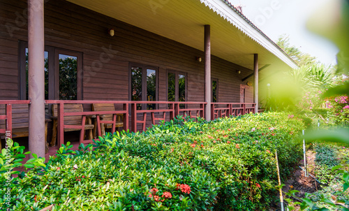 Wooden Resort Bungalow Exterior with Tropical Garden View. A serene exterior view of a wooden resort lodge with a private veranda, nestled beside a vibrant green tropical garden with red flowers.