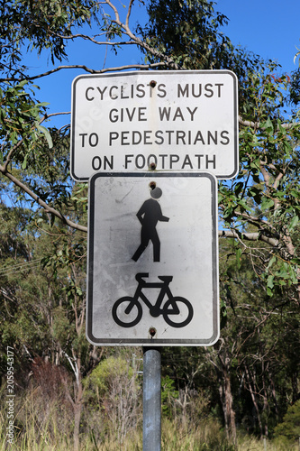 Regulatory road sign stating cyclists must give way to pedestrians on footpath with matching icons