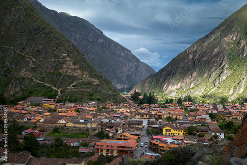 Obraz na płótnie A colorful town spreads across the floor of a steep Andean valley surrounded by green mountains and dramatic clouds in Ollantaytambo, Peru