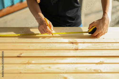 Female carpenter's hands measuring a wooden plank with a pencil and a yellow tape measure.