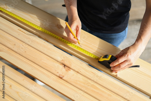 Female carpenter's hands measuring a wooden plank with a yellow tape measure.