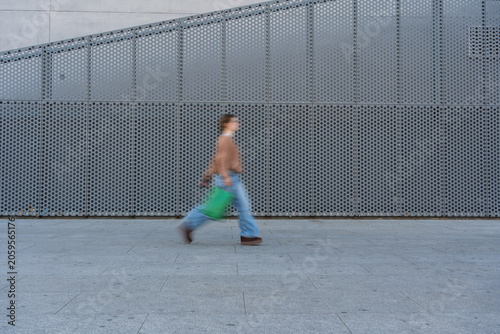 Принти на полотні Person walking quickly past a modern perforated wall carrying a green shopping b