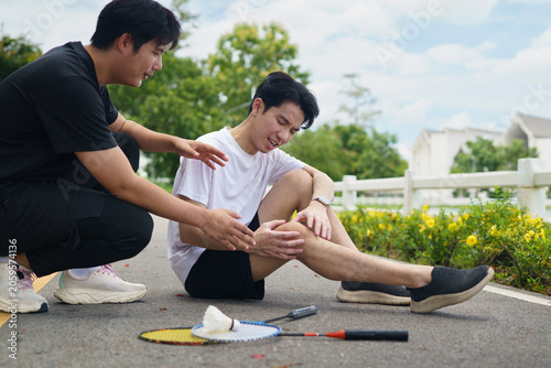 Asian man suffering knee injury during badminton game while friend helps outdoors