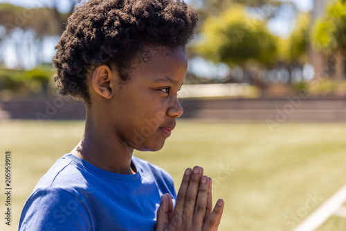 African American schoolboy standing on field near white line facing right, wearing blue tee praying