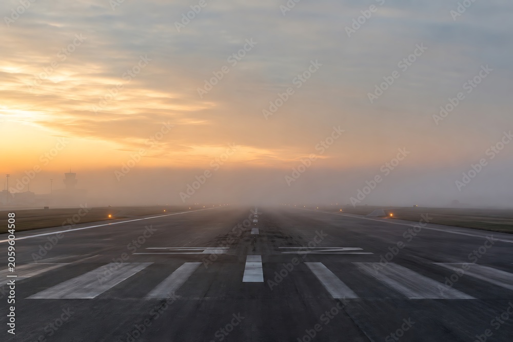 Fototapeta premium A misty sunrise over an empty airport runway with runway lights visible through the fog.
