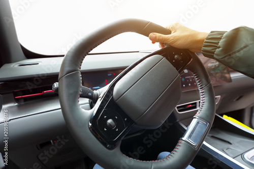 Close up of a driver hand holding a modern leather steering wheel with red stitching inside a car.