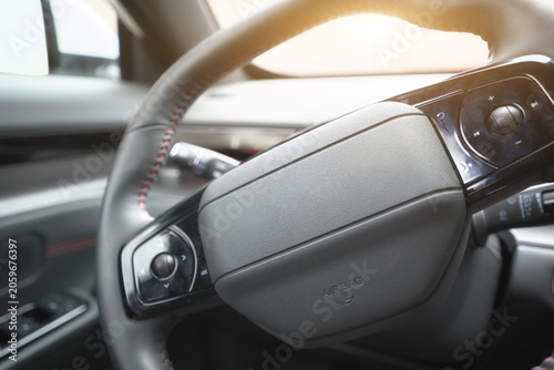 Close up view of a modern car steering wheel with leather trim and red stitching under soft sunlight.