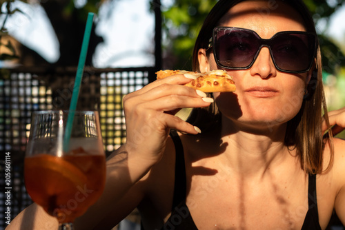 Obraz na plátne Young woman in sunglasses sholding a slice of pizza in her hand and  taking a bite out of it
