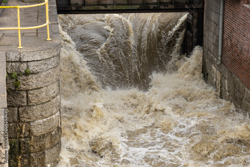 Rushing muddy river water gushing down an urban dam and concrete lock system gate with foam and splashes during a city flood. Hydroelectric infrastructure, deluge and safety risk