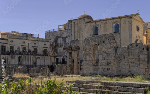 Ancient ruins of the Temple of Apollo with the San Paolo Apostolo church background in Ortigia, Syracuse, Italy. Historic archaeological site and old town buildings in Sicily.