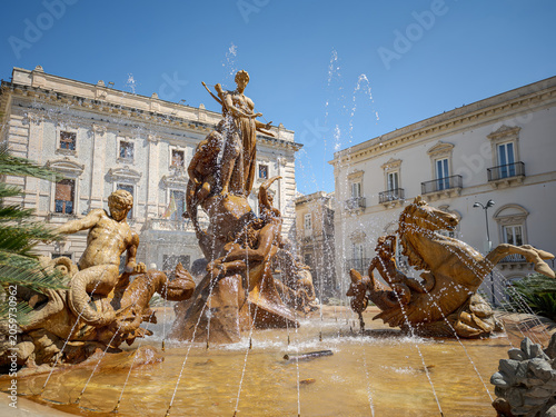 Wide angle view of the famous mythological fountain in Siracusa, Italy. Elegant urban public square featuring ornate classical architecture and deep blue summer sky.