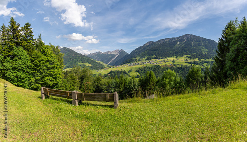 park bench by the road side with view to the mountains from great valley of Walser and a small village, scattered settlement on the steep slope with alpine farmhouses, flowered meadows and forests