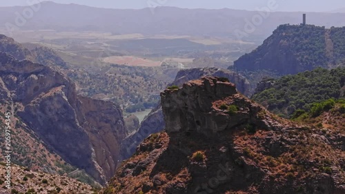 Drone footage ascending sideways past a weathered summit boulder, framing the sheer gorge drop, glinting reservoir below, distant plains, and a lone hilltop tower under summer haze.
