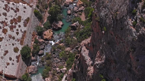 Drone footage ascending forward over El Chorro's churning river, framed by towering cliff walls and dam stonework, with turquoise pools, boulders, and a cliff-hugging path below.