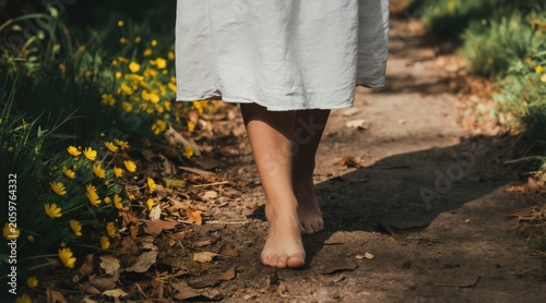 Woman walks barefoot on a path surrounded by yellow flowers and green grass. Her white dress flows as she moves through nature enjoying a peaceful moment.