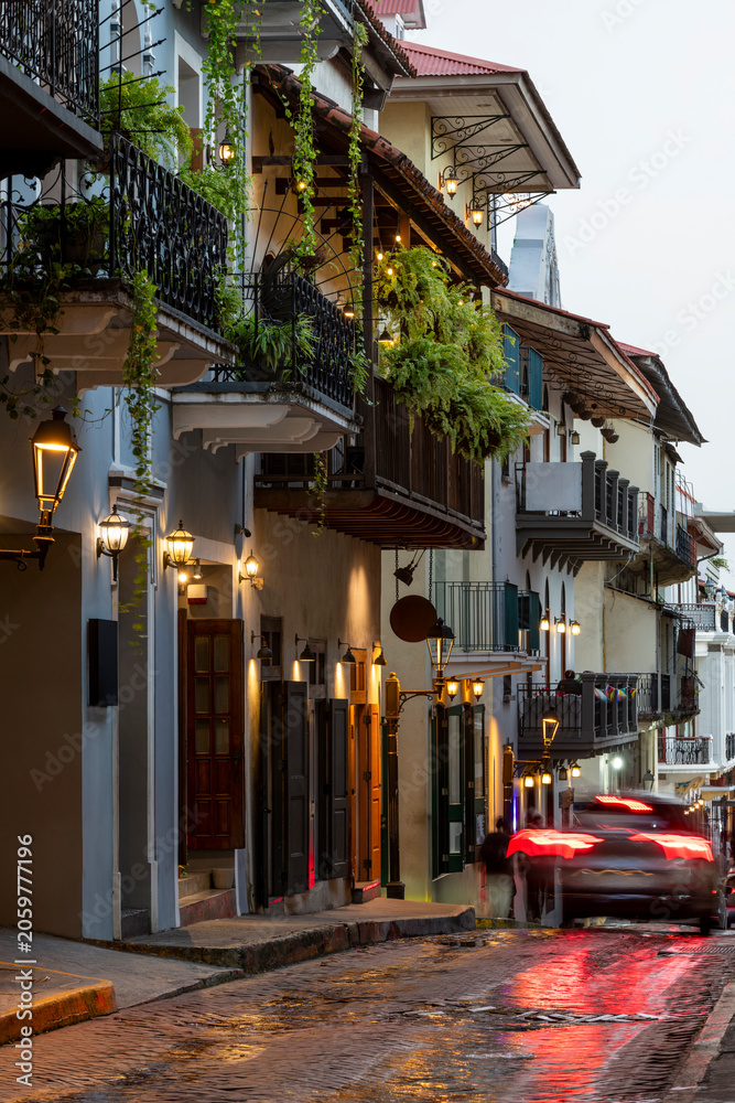 Obraz premium Colonial architecture lines a cobbled street in Casco Viejo, Panama city, illuminated by charming streetlights and vehicle tail lights reflecting on the wet pavement - stock photo