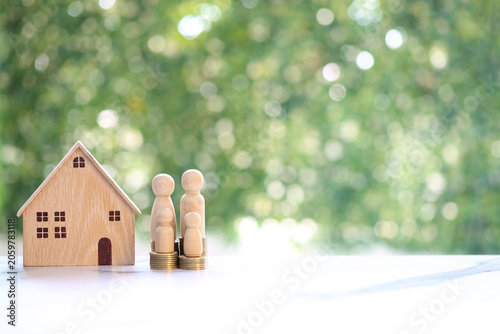 Wooden house model and a family of four wooden figures standing on stacks of coins against a soft green bokeh background, representing financial planning for a family home.