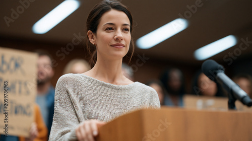 A woman in her forties speaks at a city council podium, a small group of supporters behind her holding handwritten signs advocating for labor rights and legal protections, the