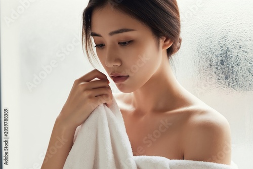 Young Asian woman in towel after shower. She has clear skin and dark hair. Woman looks thoughtful near steamy window. She feels fresh and relaxed.