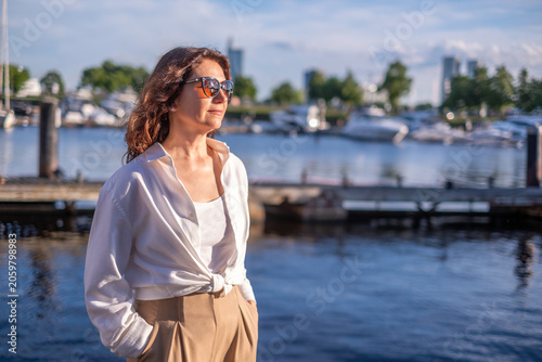 Vital brunette woman over 50 looking toward sunset at marina, enjoying peaceful golden hour and elegant vacation lifestyle with copy space
