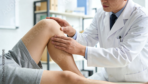 Palpating doctor in white coat, tie bracing patient's knee on exam table, patient in gray shorts