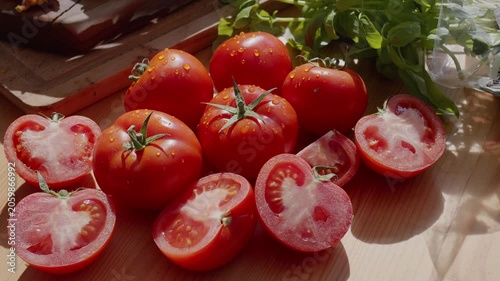 Slow camera movement along ripe tomatoes and cut tomatoes on a sunlit wooden table with greenery.
