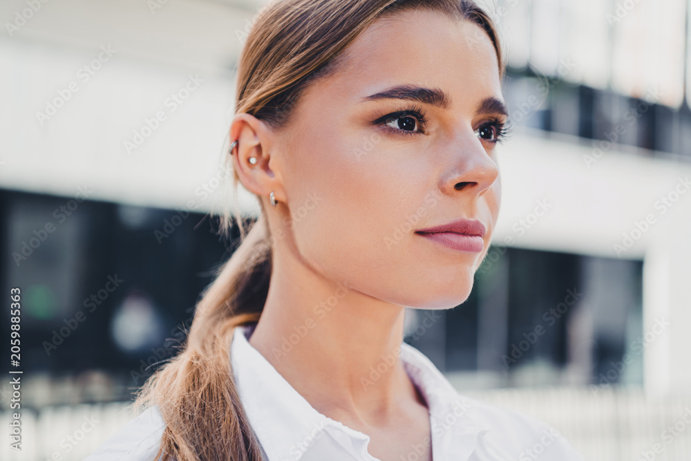 Fototapeta premium Confident young professional woman portrait outdoors looking thoughtful with ponytail, natural makeup and ear piercings