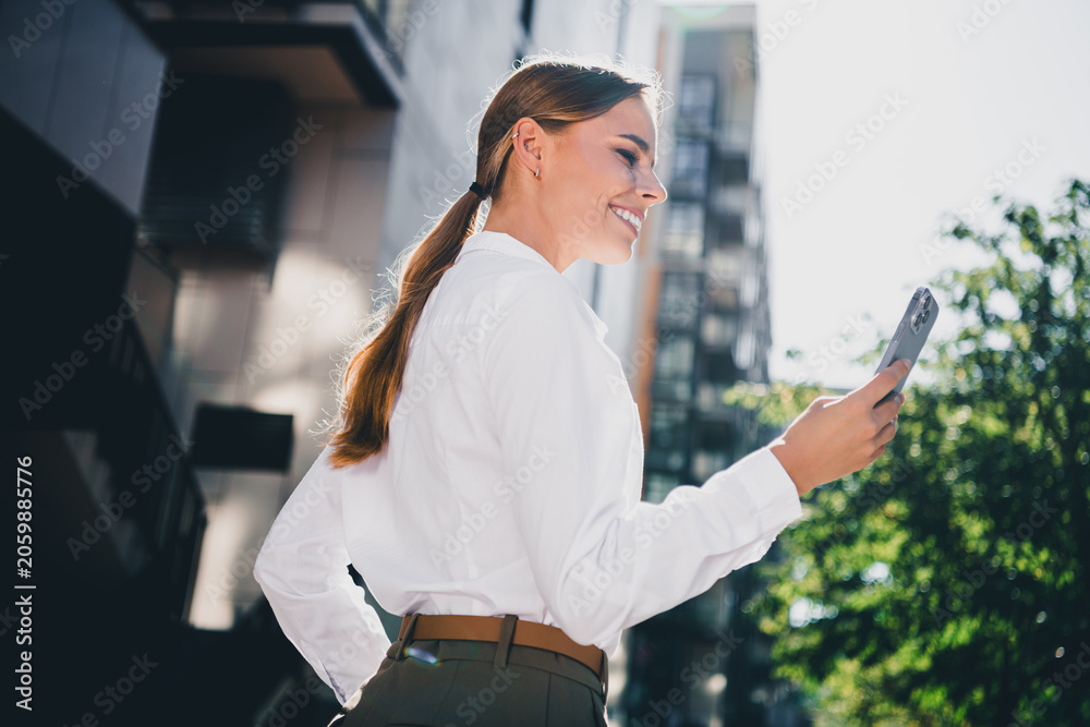 Fototapeta premium Smiling businesswoman using smartphone outdoors in an urban setting, confident professional checking her phone on a sunny city street for work and lifestyle