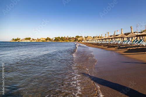 Empty sand beach with straw umbrella and lounge row along water surf line at morning. Calm sea near old town peninsula under clear blue sky, summer holiday resort, Side, Turkey.