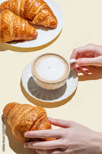 Hands holding a cup of cappuccino coffee and croissant on a yellow isolated background