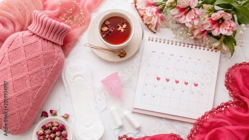 Flat lay of period care essentials with calendar, menstrual cup, pad, tampons, hot water bottle, tea, flowers and pink lingerie for cycle comfort