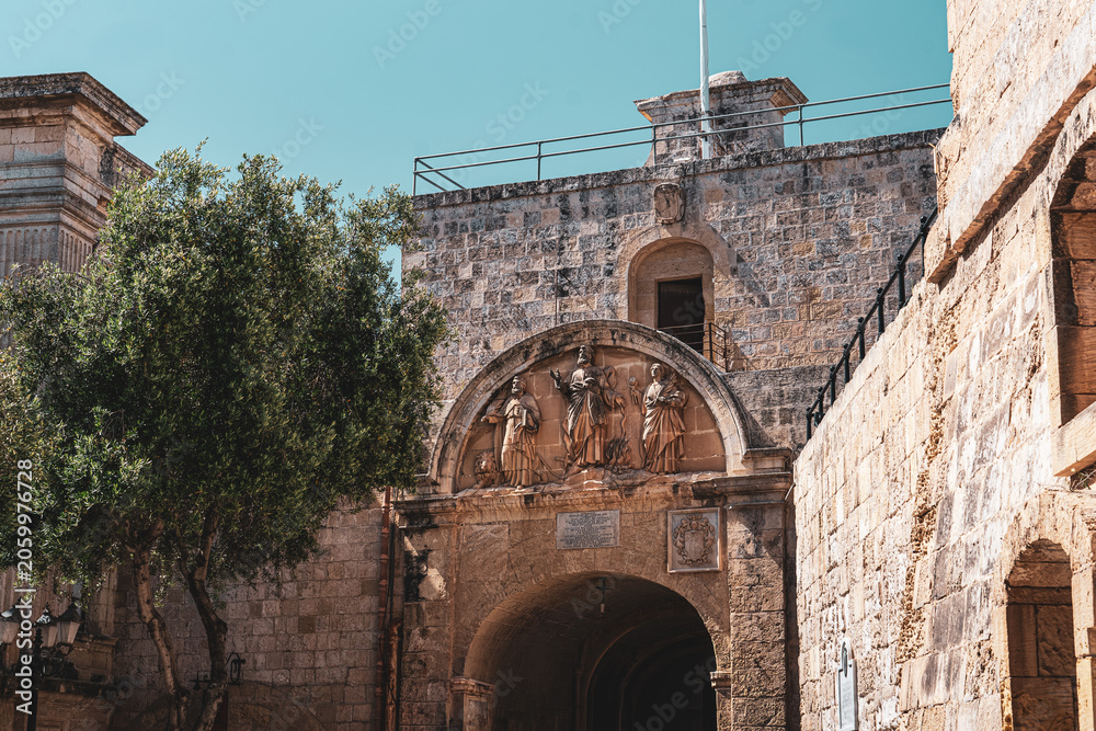 Obraz premium view from behind mdina gate, malta with limestone brick and detailed statues above the archway with clear blue skies overhead in summer sunlight and green lush tree to the left