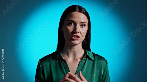 Woman speaking to camera against blue background. Caucasian female presenter gesturing while talking. Professional vlog or interview setup for communication concept.