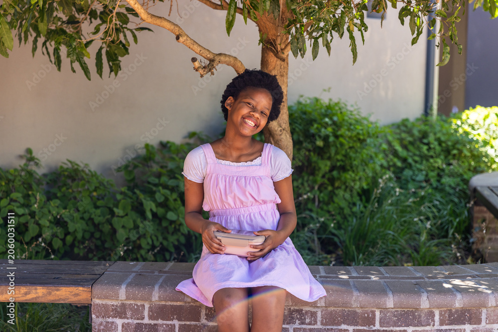 Naklejka premium African American child wearing pink dress sitting on brick bench under tree cradling bowl smiling
