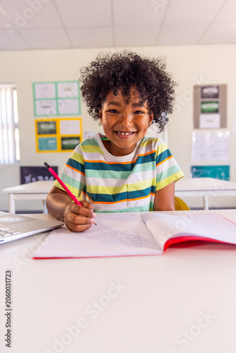 African American child in striped tee holding red pencil at school desk, smiling over red notebook