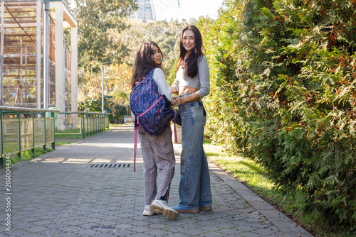 Two young women standing on a path near trees at a park on a sunny day enjoying their time together while wearing casual clothing