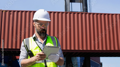 Serious worker utilizes tablet for container inspection during quality control assessment within busy industrial shipping yard, wearing hard hat, safety glasses, and work gloves.