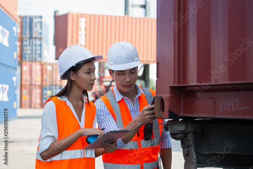 Two professionals conduct detailed inspection of shipping container within busy port environment while wearing safety vests and hard hats, emphasizing quality control processes.
