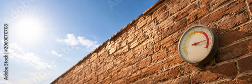 Thermometer gauge attached to a red brick exterior wall under bright blue sky and sun. Home weather monitoring.