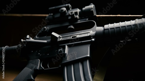 This side view displays a modular carbine rifle equipped with an optical sight and a durable sling placed on a wooden platform against a dark background, showcasing its tactical configuration