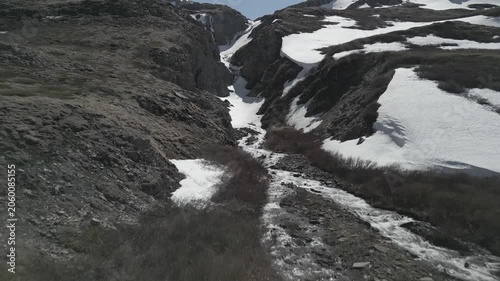 Aerial crane shot rising over scenic spring mountain waterfall with ice floes towards frozen alpine lake. Remote wilderness. RAW flat profile uncorrected video footage.